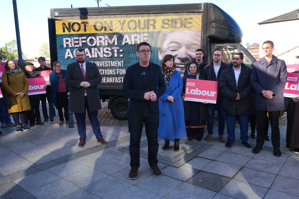Burnham speaking in front of an anti-Reform UK poster
